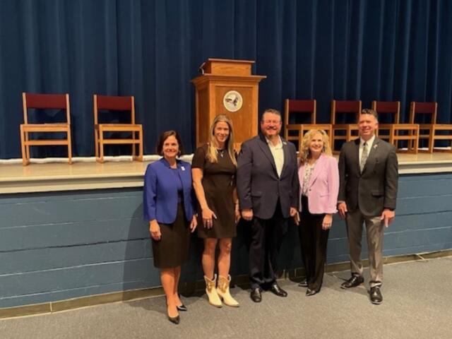 A gray concrete block base to a stage in the background, with chairs to the right and left of a podium. Well-dressed people standing together in front of the stage.