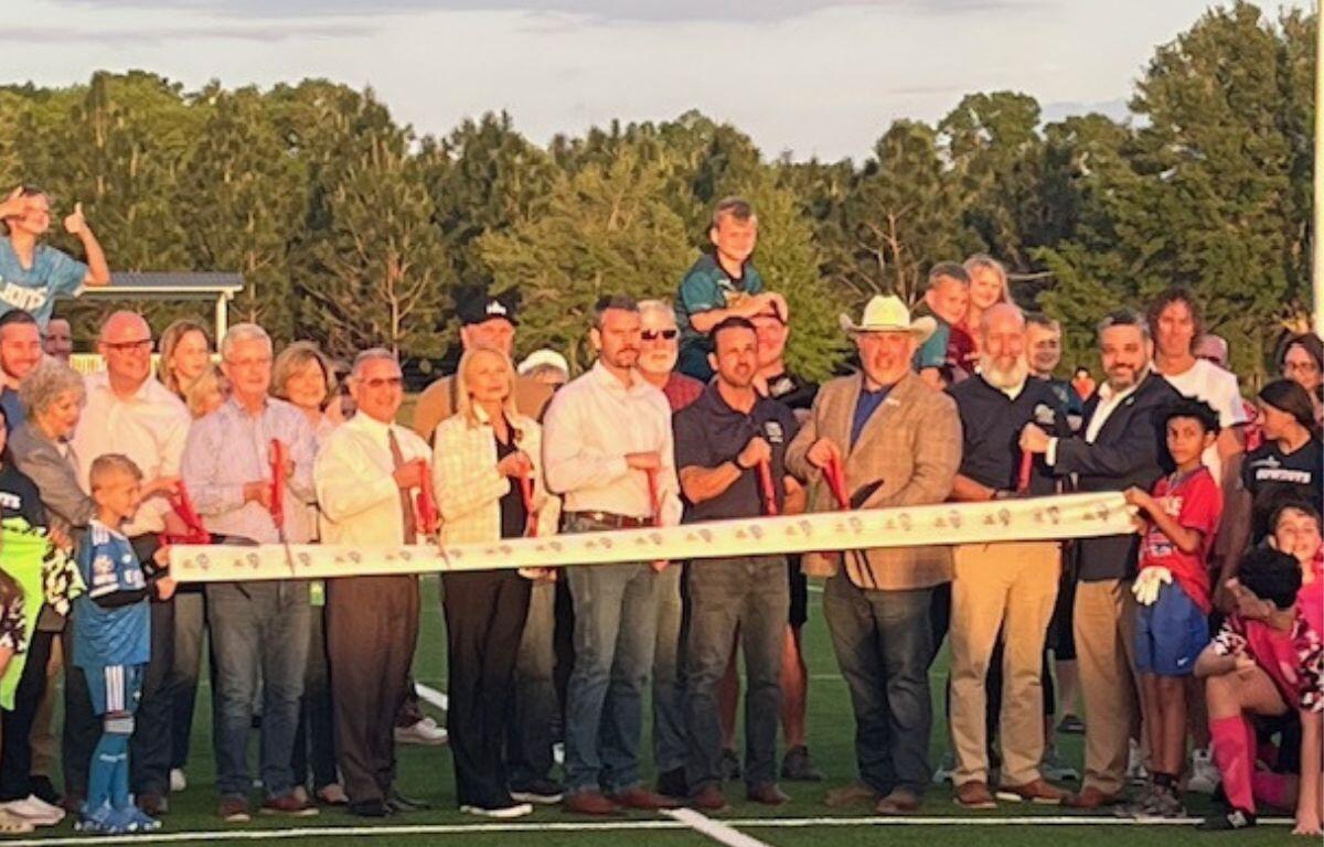 In the background a tree line, in the middle, a swell of people in rows, with the people behind the ribbon holding giant scissors. In the foreground artificial turf.
