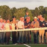 In the background a tree line, in the middle, a swell of people in rows, with the people behind the ribbon holding giant scissors. In the foreground artificial turf.