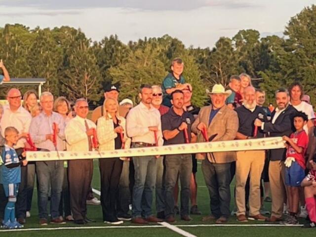 In the background a tree line, in the middle, a swell of people in rows, with the people behind the ribbon holding giant scissors. In the foreground artificial turf.