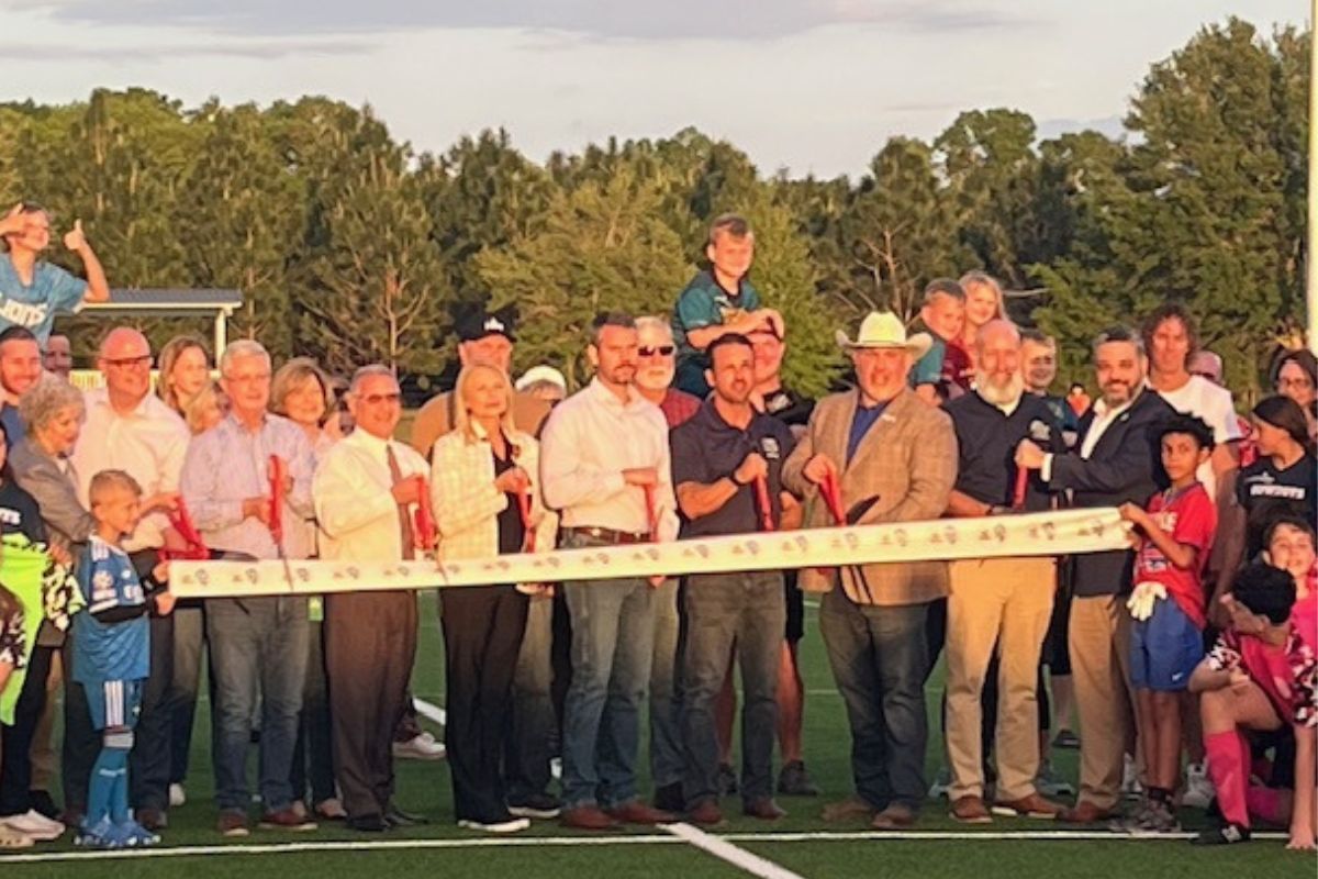 In the background a tree line, in the middle, a swell of people in rows, with the people behind the ribbon holding giant scissors. In the foreground artificial turf.
