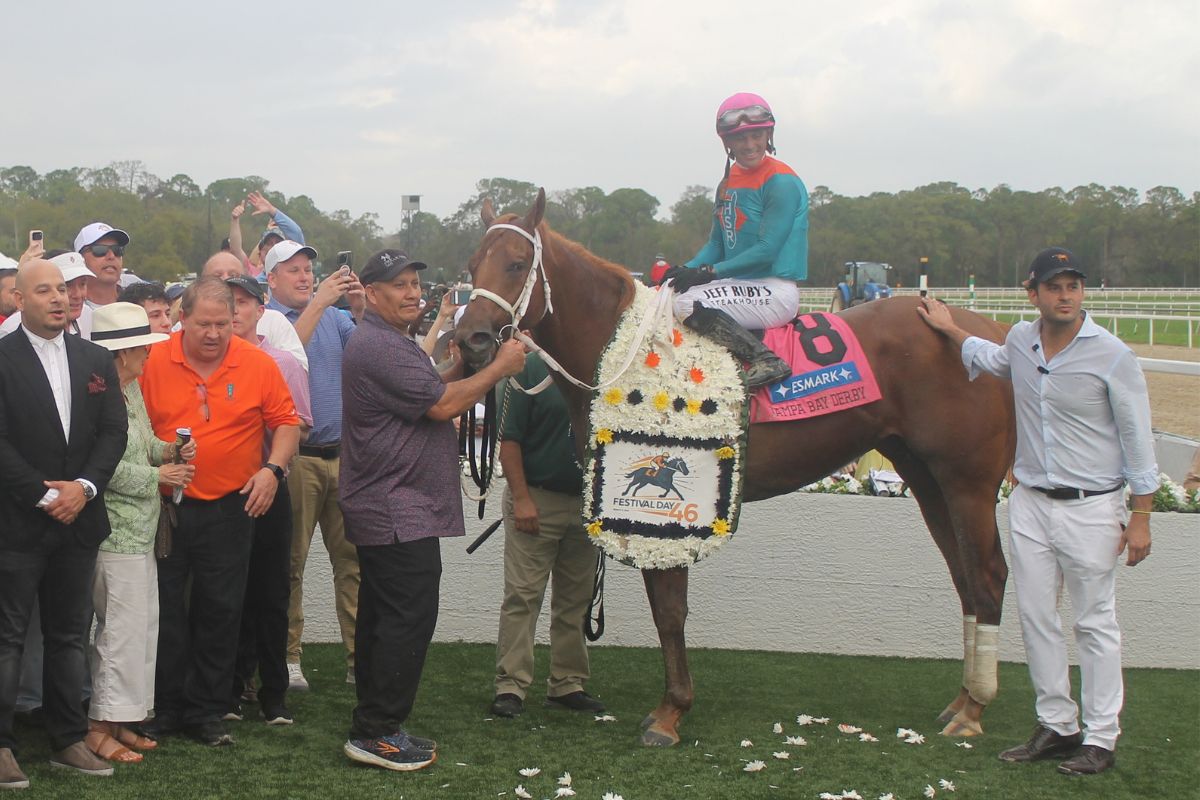 People standing next to a horse, with a garland of flowers wrapped over the horse, and rider, wearing a helmet and gallops, with his legs in the irons over a saddle cloth, with a man with his hand on the horse's flank to the extreme right.