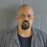 A colored image of a bald man with a goatee posed in front of a grey wall.