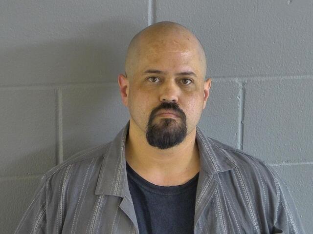 A colored image of a bald man with a goatee posed in front of a grey wall.