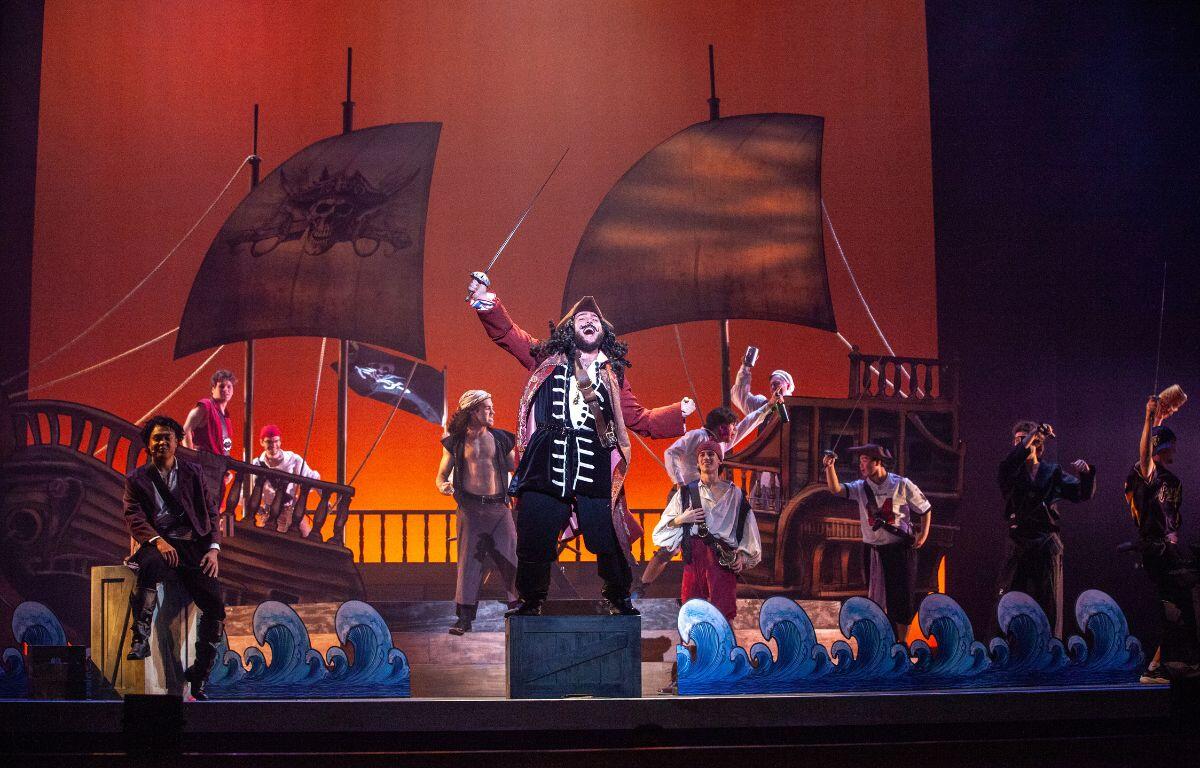 A colorful photo with a red backdrop. A pirate ship set is in the background with a group of pirates in the foreground.