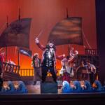 A colorful photo with a red backdrop. A pirate ship set is in the background with a group of pirates in the foreground.