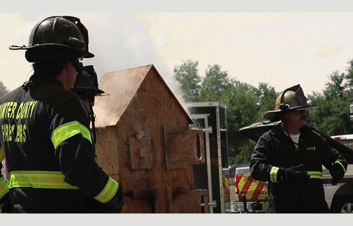 Two Sumter County firefighters putting on a demonstration by a miniature wooden house.