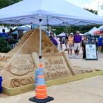A sand sculpture advertising the Sand and Sun Festival underneath a white tent.