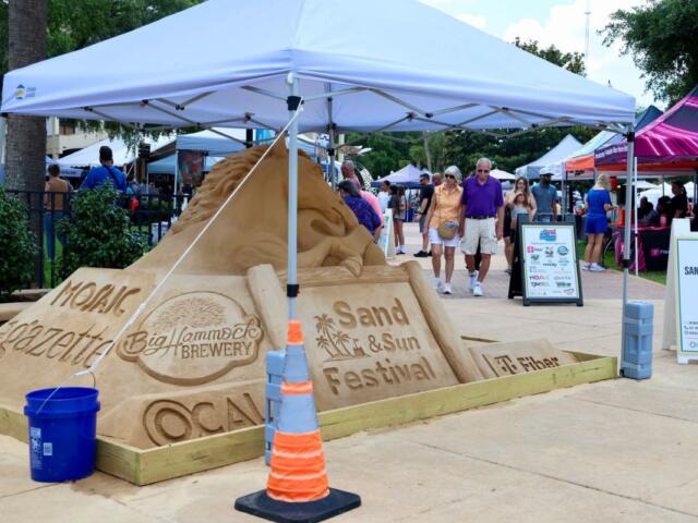 A sand sculpture advertising the Sand and Sun Festival underneath a white tent.