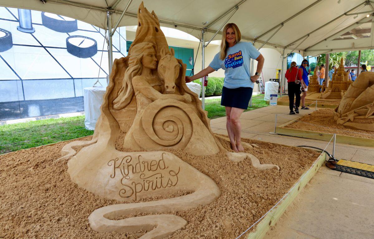 Woman posing next to a large sand sculpture titled 'Kindred Spirits' under a festival tent.