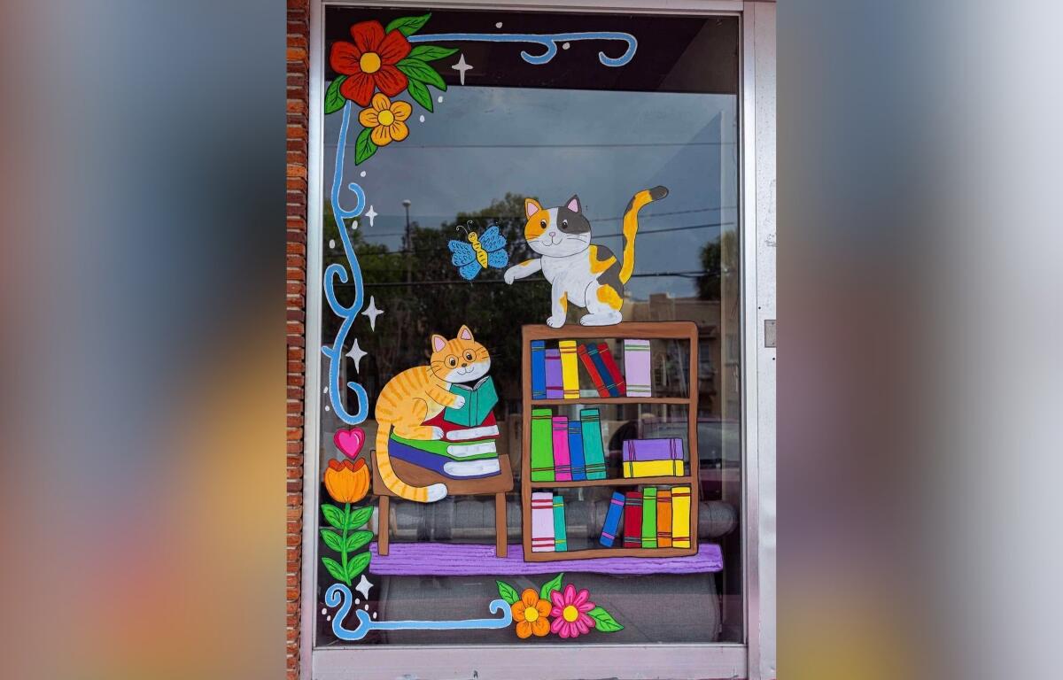 Colorful painted window mural of two cats with a bookshelf and flowers on a storefront display.