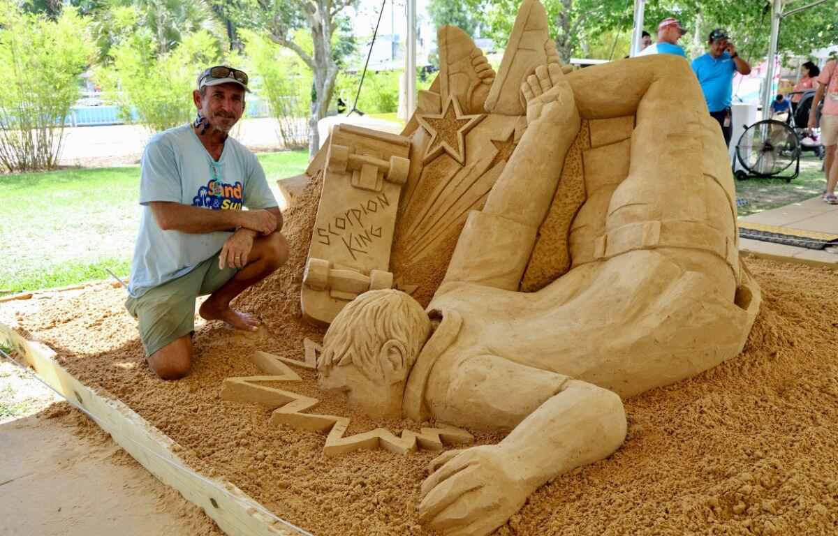 Sand sculpture of a reclining figure with star shapes and geometric blocks, posed beside the artist kneeling in a light blue shirt under a canopy at a sunny park event.