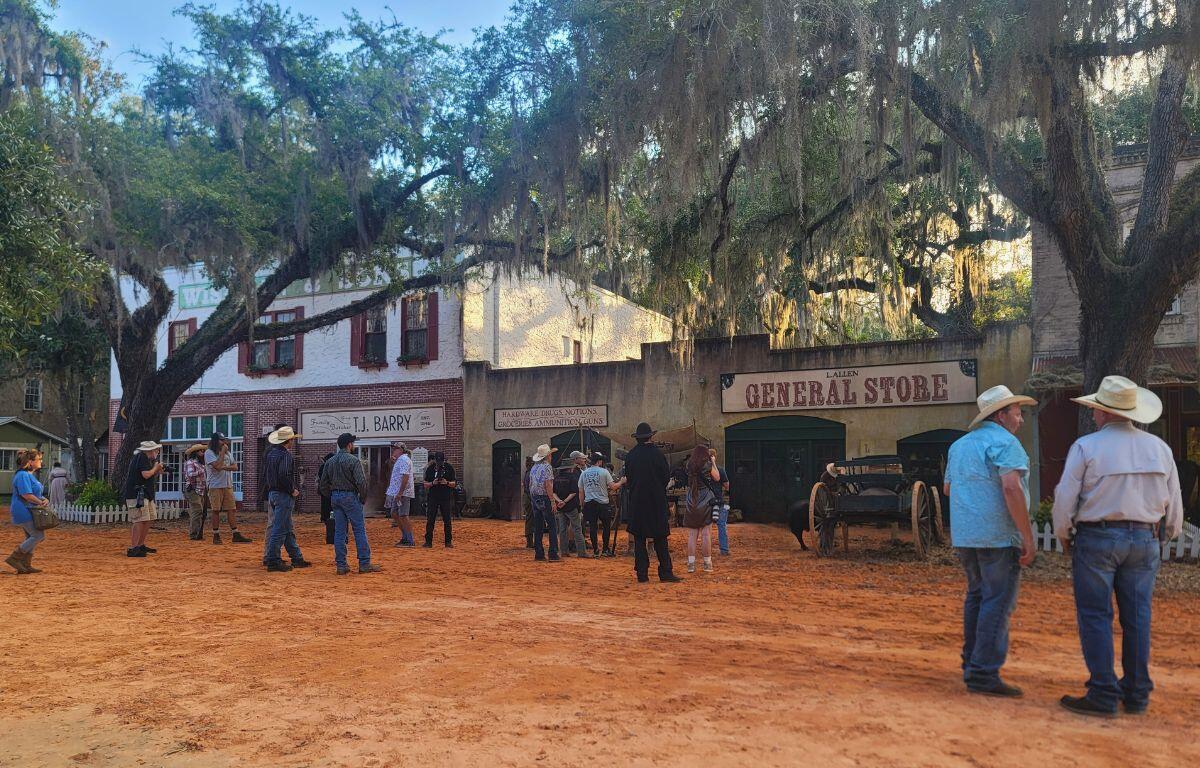 A town is set to look like it's from the 1800s. Men in cowboy hats stand around the area, talking while standing on a red, clay road.