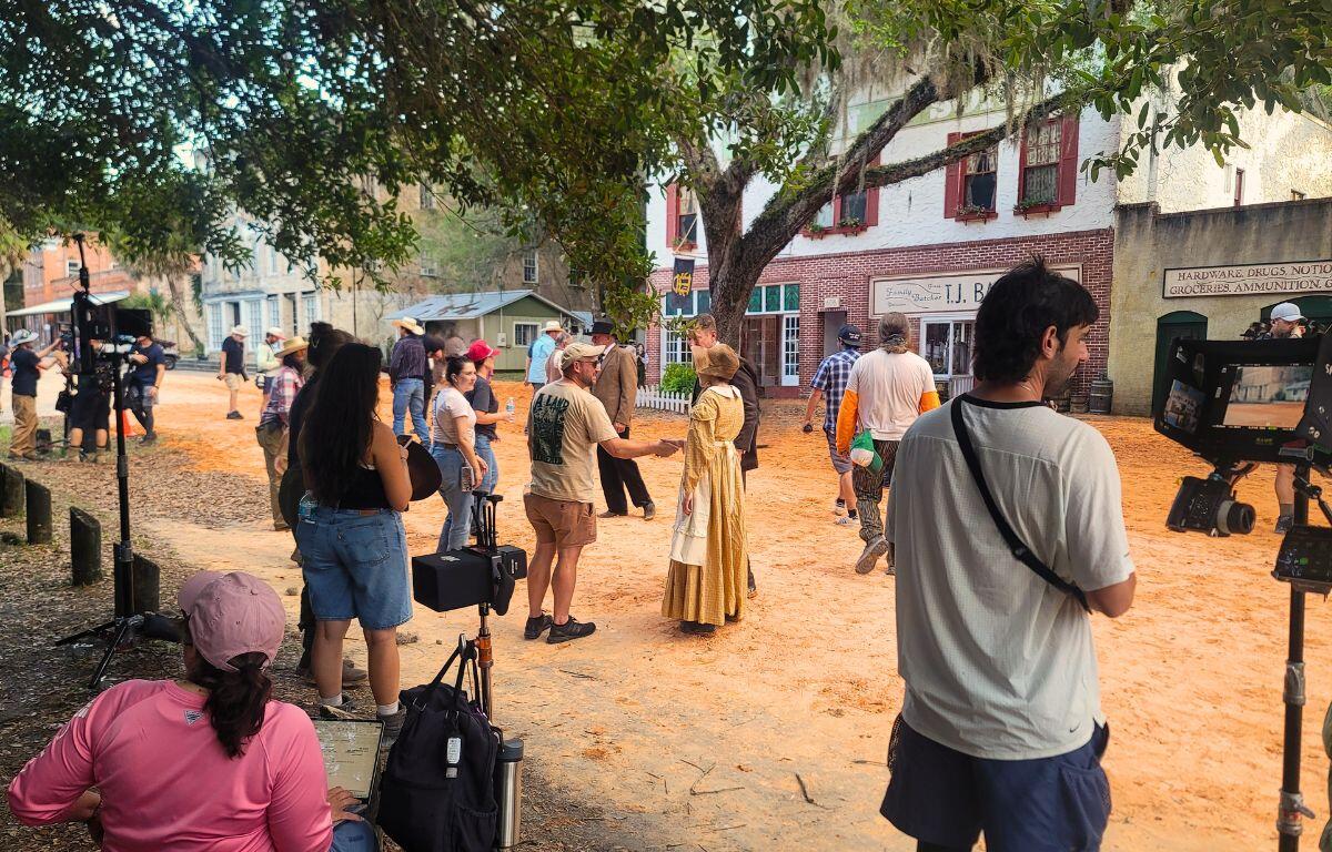 A production crew stands around a town area decorated to look like the 1800s. Cameras and various film tools are around the area while actors and crew talk and walk around.