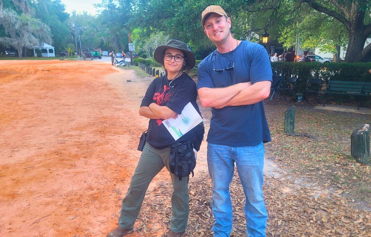 Two people stand next to each other with on a red, clay road with a town behind them.