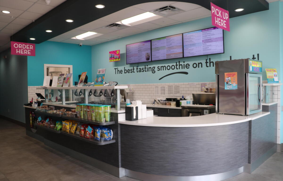 Bright smoothie shop counter with pink 'Order Here' and 'Pick Up Here' signs, overhead menu boards, and snack display along a curved counter.