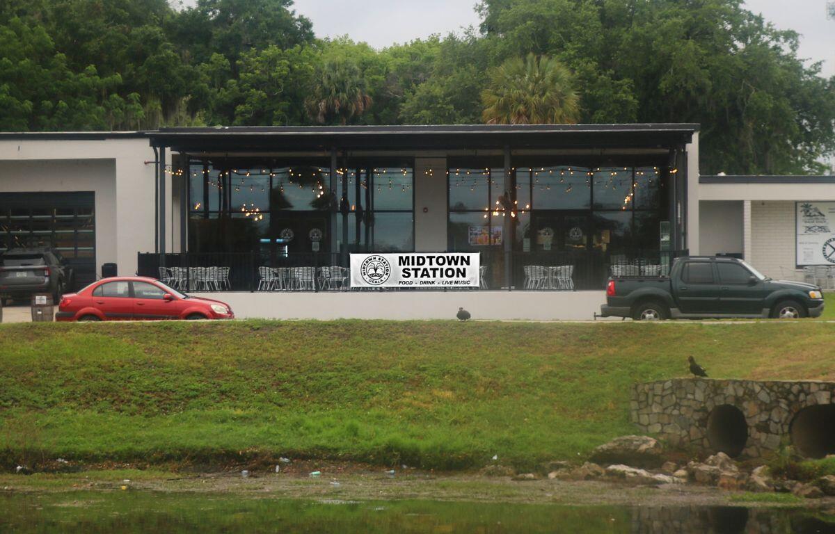 The exterior of a restaurant has two cars parallel parked in front of it. A large banner hanging from a fence surrounding an outdoor patio of the restaurant reads, "Midtown Station."