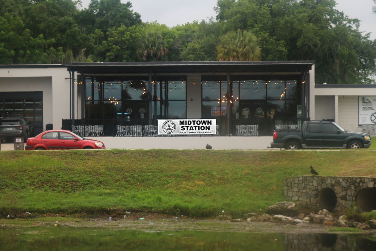 The exterior of a restaurant has two cars parallel parked in front of it. A large banner hanging from a fence surrounding an outdoor patio of the restaurant reads, "Midtown Station."