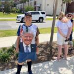 A woman wearing a long shirt with a print of President Donald J. Trump on the front. there are people standing to her right. She's standing on a sidewalk, with curb appeal to her back, and vehicles and a building behind her to her left.