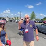 A woman wearing a patriotic hat, holding hats in her left hand and a man standing lot with cars to the left and right, with light poles and trees in the background.