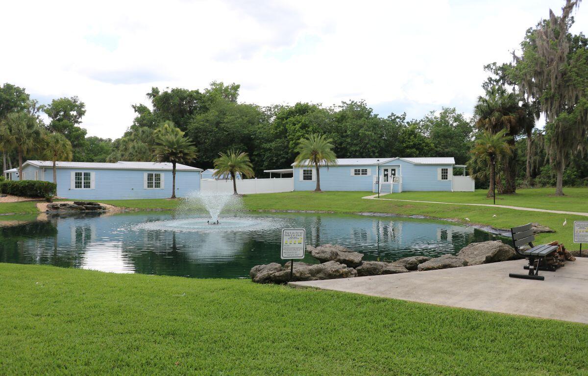 Two blue building sit in the distance with a large pond in front of them. A concrete patio with a bench sits along the pond.