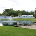 Two blue building sit in the distance with a large pond in front of them. A concrete patio with a bench sits along the pond.
