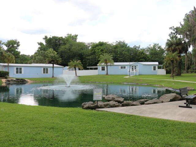 Two blue building sit in the distance with a large pond in front of them. A concrete patio with a bench sits along the pond.