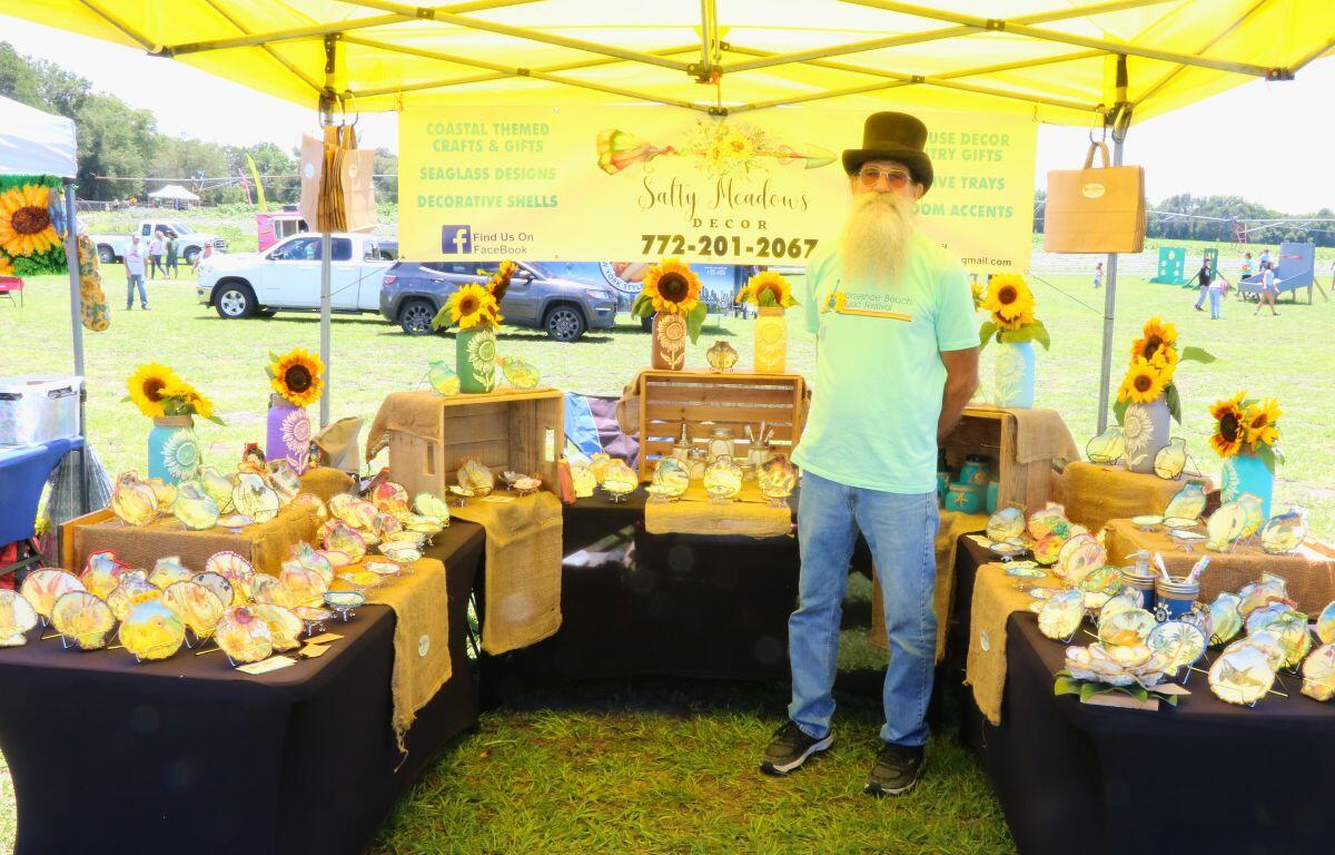 A man with a top hat stands under a yellow canopy at a market. Tables with black tablecloths surround him with decorative shells and sunflowers scattered on them.