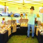 A man with a top hat stands under a yellow canopy at a market. Tables with black tablecloths surround him with decorative shells and sunflowers scattered on them.