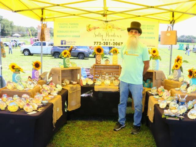 A man with a top hat stands under a yellow canopy at a market. Tables with black tablecloths surround him with decorative shells and sunflowers scattered on them.