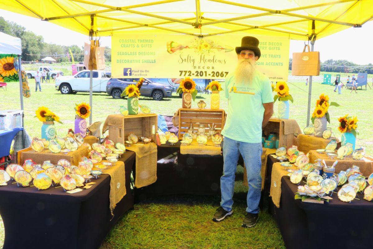 A man with a top hat stands under a yellow canopy at a market. Tables with black tablecloths surround him with decorative shells and sunflowers scattered on them.