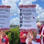 Signs with writing festooned with garland, being held by people on the left and right, with shrubbery in the background.