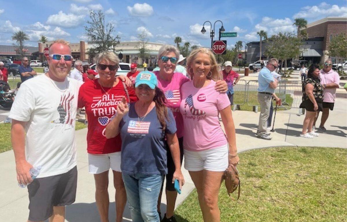 A man on the left and four women stand together, with grass and a sidewalk in the background, with a swell of people, trees and street signs.