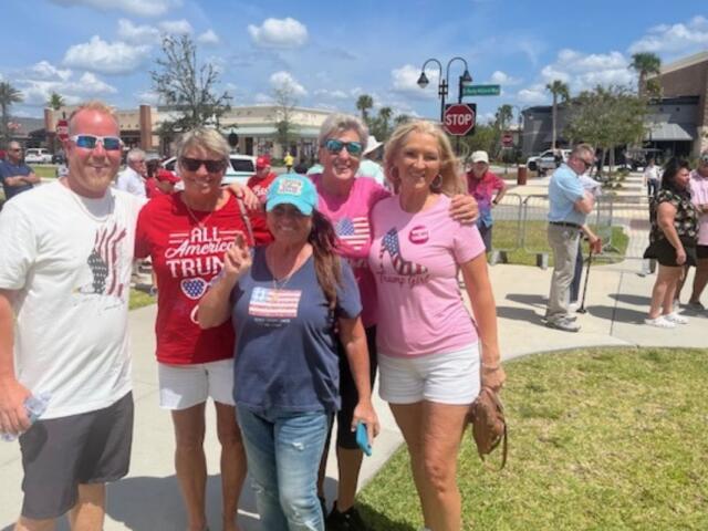 A man on the left and four women stand together, with grass and a sidewalk in the background, with a swell of people, trees and street signs.