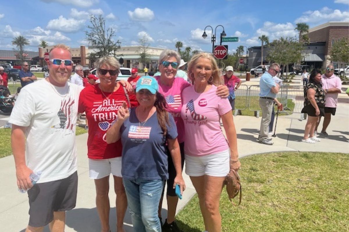 A man on the left and four women stand together, with grass and a sidewalk in the background, with a swell of people, trees and street signs.
