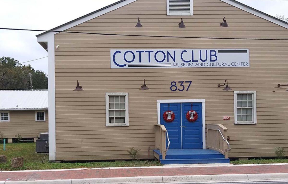 Beige wooden building with blue double doors, wreaths, and a sign reading COTTON CLUB Museum and Cultural Center; address 837.