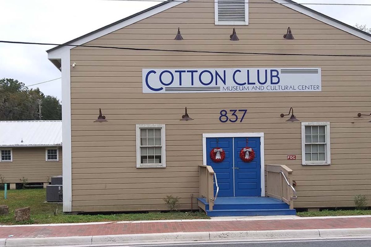 Beige wooden building with blue double doors, wreaths, and a sign reading COTTON CLUB Museum and Cultural Center; address 837.