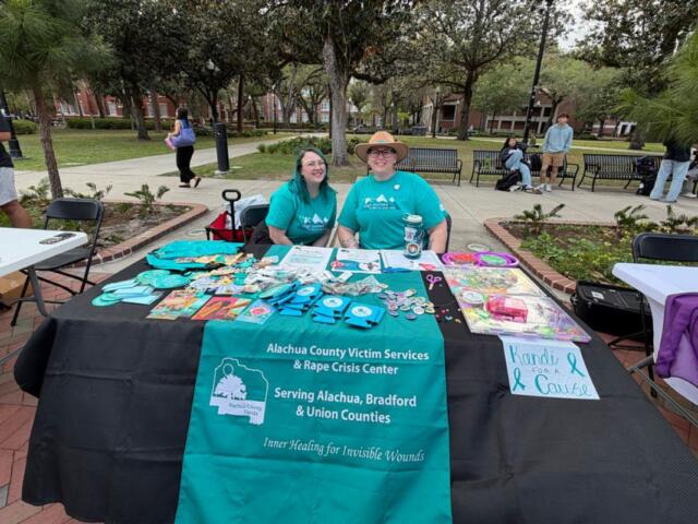 A colored image of two people in light blue shirts smiling behind a booth.