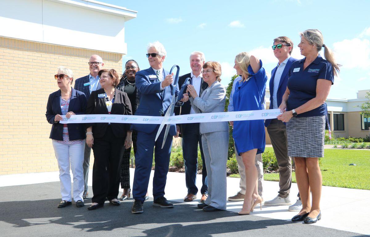 A colored photo of a group of people dressed in business casual posing before cutting a ribbon.