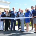 A colored photo of a group of people dressed in business casual posing before cutting a ribbon.