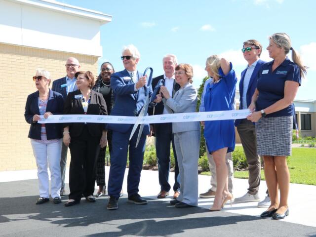 A colored photo of a group of people dressed in business casual posing before cutting a ribbon.