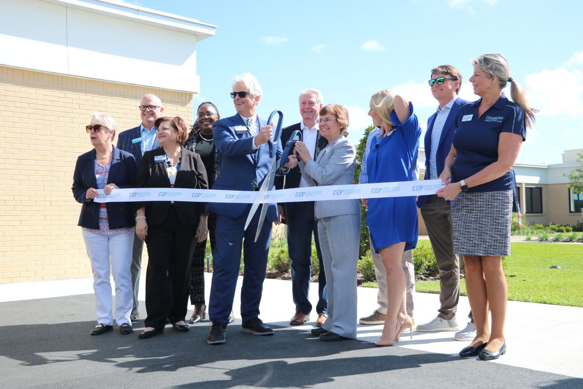 A colored photo of a group of people dressed in business casual posing before cutting a ribbon.