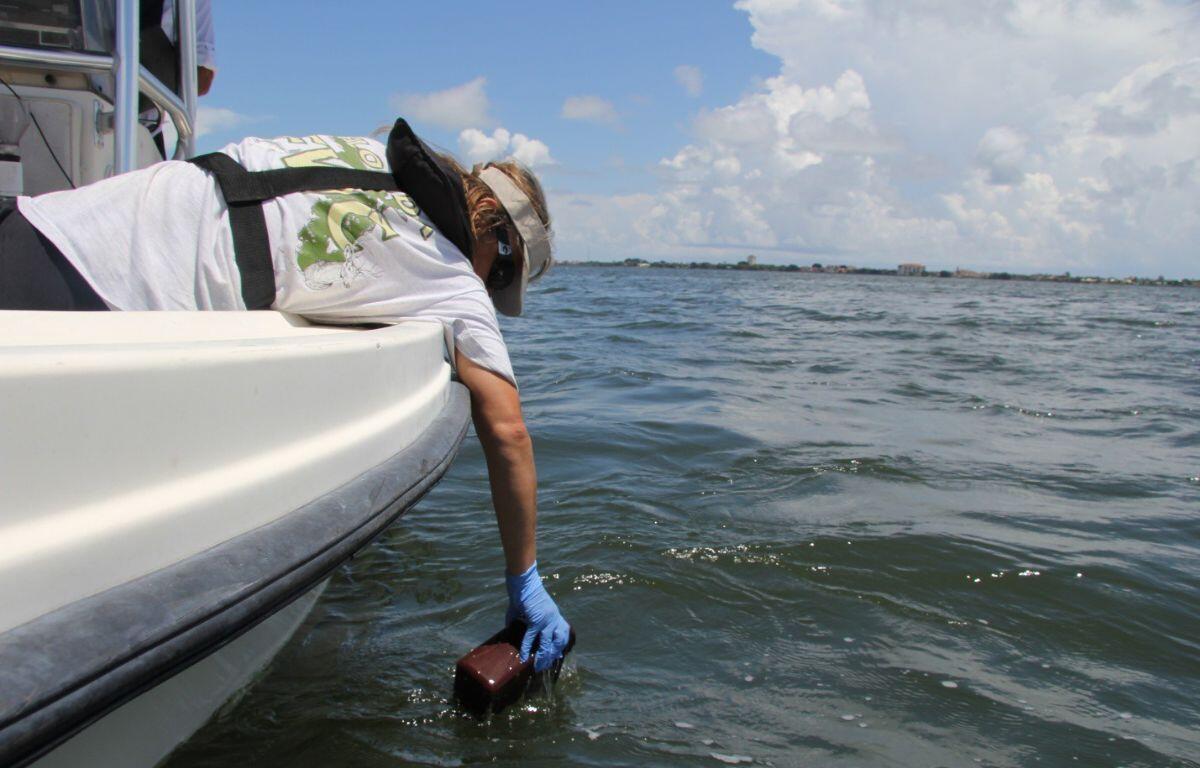 Crew member on a boat reaches into the water with a gloved hand, holding a small brown rectangular device near the surface.
