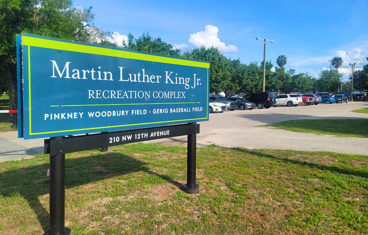 Blue sign reads 'Martin Luther King Jr. Recreation Complex' with 'Pinkney Woodbury Field · Gerig Baseball Field' and address 210 NW 12th Avenue; a parking lot and trees are in the background. Cars take up all available parking spots.