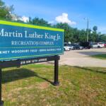 Blue sign reads 'Martin Luther King Jr. Recreation Complex' with 'Pinkney Woodbury Field · Gerig Baseball Field' and address 210 NW 12th Avenue; a parking lot and trees are in the background. Cars take up all available parking spots.