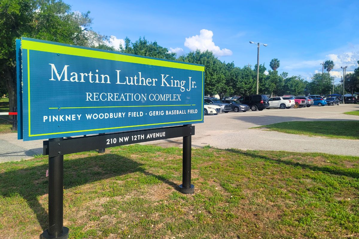 Blue sign reads 'Martin Luther King Jr. Recreation Complex' with 'Pinkney Woodbury Field · Gerig Baseball Field' and address 210 NW 12th Avenue; a parking lot and trees are in the background. Cars take up all available parking spots.