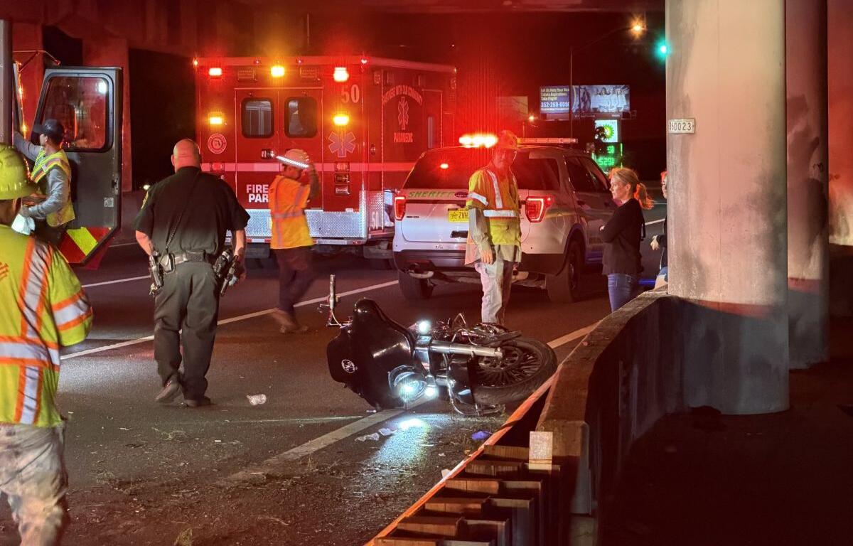 Motorcycle on its side in the road at night as firefighters, police, and a responding ambulance handle a crash under an overpass with flashing lights nearby
