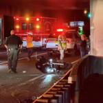 Motorcycle on its side in the road at night as firefighters, police, and a responding ambulance handle a crash under an overpass with flashing lights nearby