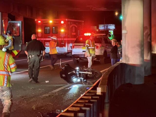 Motorcycle on its side in the road at night as firefighters, police, and a responding ambulance handle a crash under an overpass with flashing lights nearby