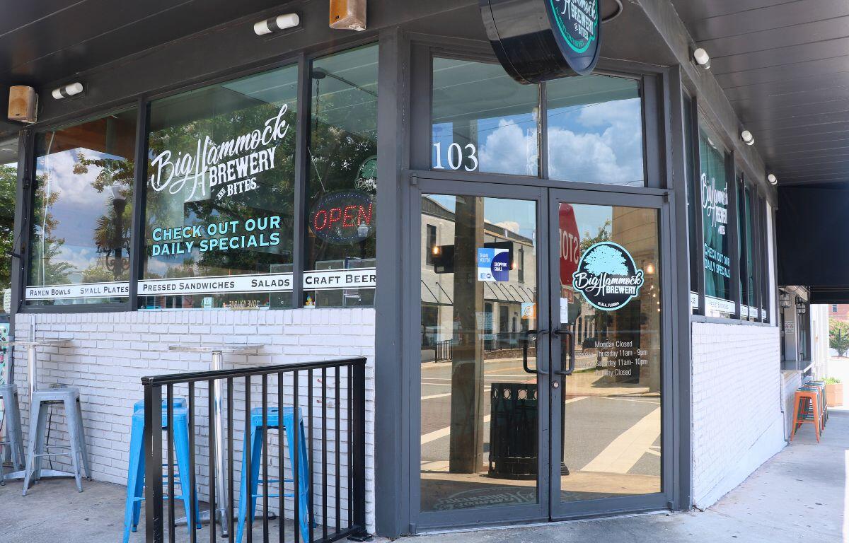 The exterior of a restaurant features signs reading, "Big Hammock Brewery & Bites." Outdoor seating lines the walls on a sidewalk with a small, black fence next to the entrance.